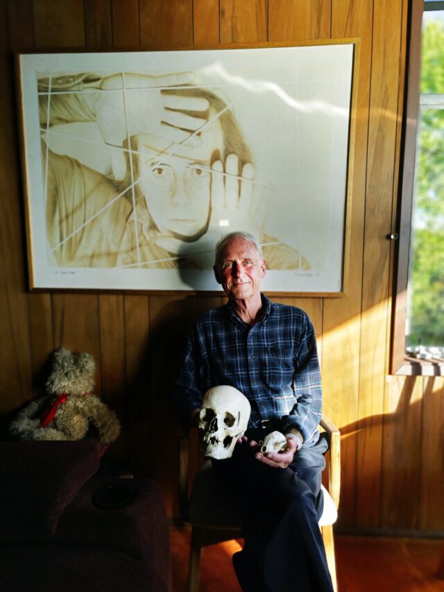 older white man in plaid shirt sitting under art work with a skull on his lap