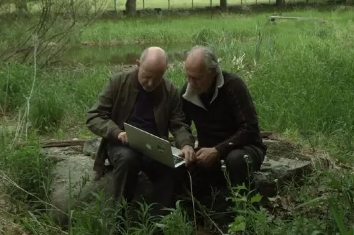 Werner Herzog and Rafael Yuste in a field sitting on a log while looking at a laptop screen