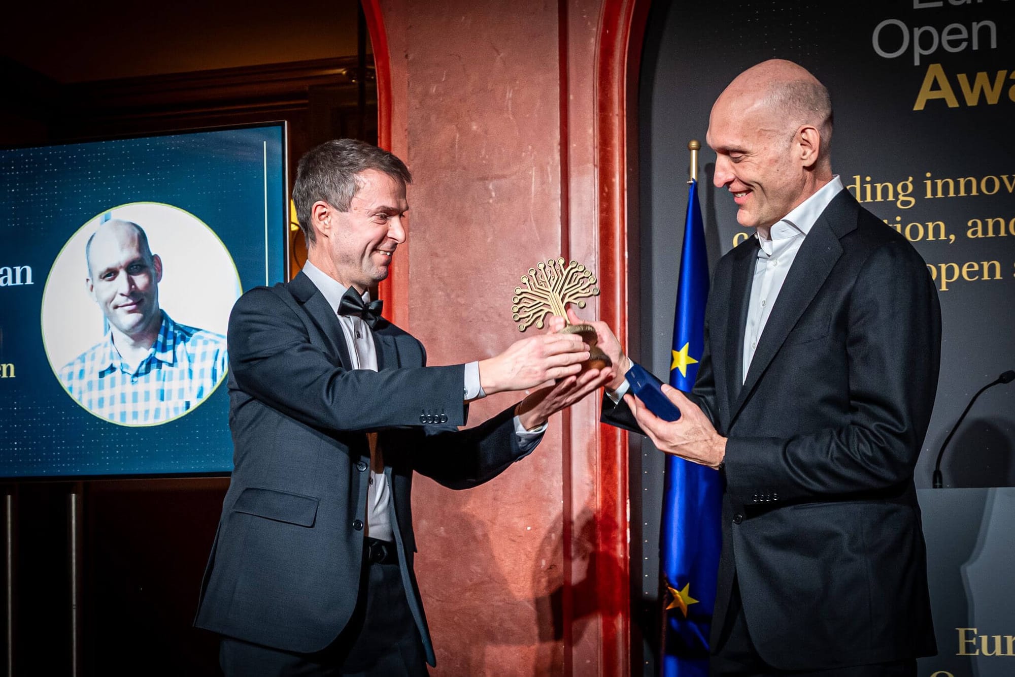 this picture shows two people in the frame, on the left is daniel wearing a light blue suit, handing over a tree-themed award to greg on the right, who is wearing a black colored suit